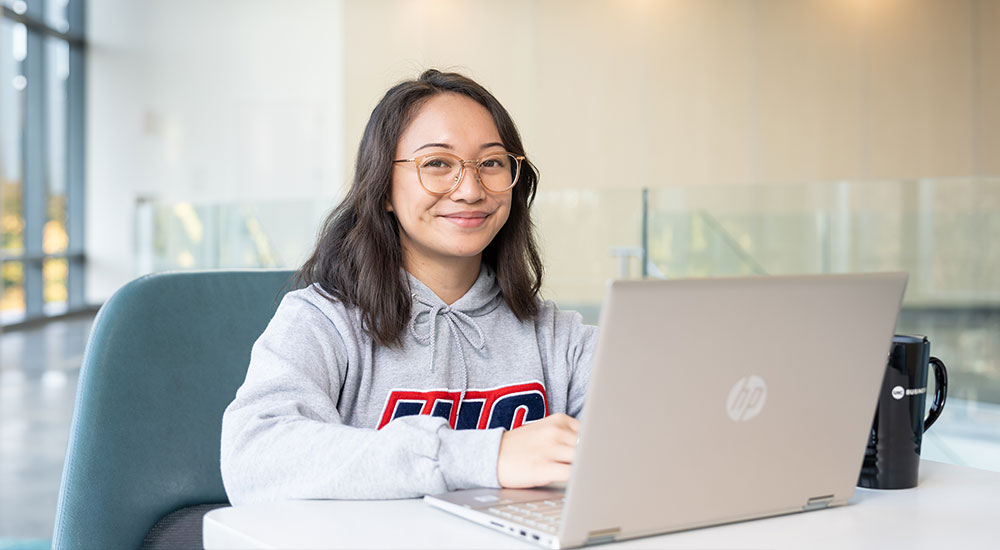 UIC mba student smiling in a university sweatshirt working on a laptop in a bright, modern study space.
